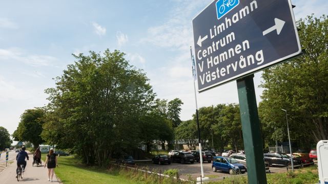Cykelvägen i Limhamn med träd och grönska runt om. I högra hörnet en skylt som visar vilket håll Limhamn ligger åt 